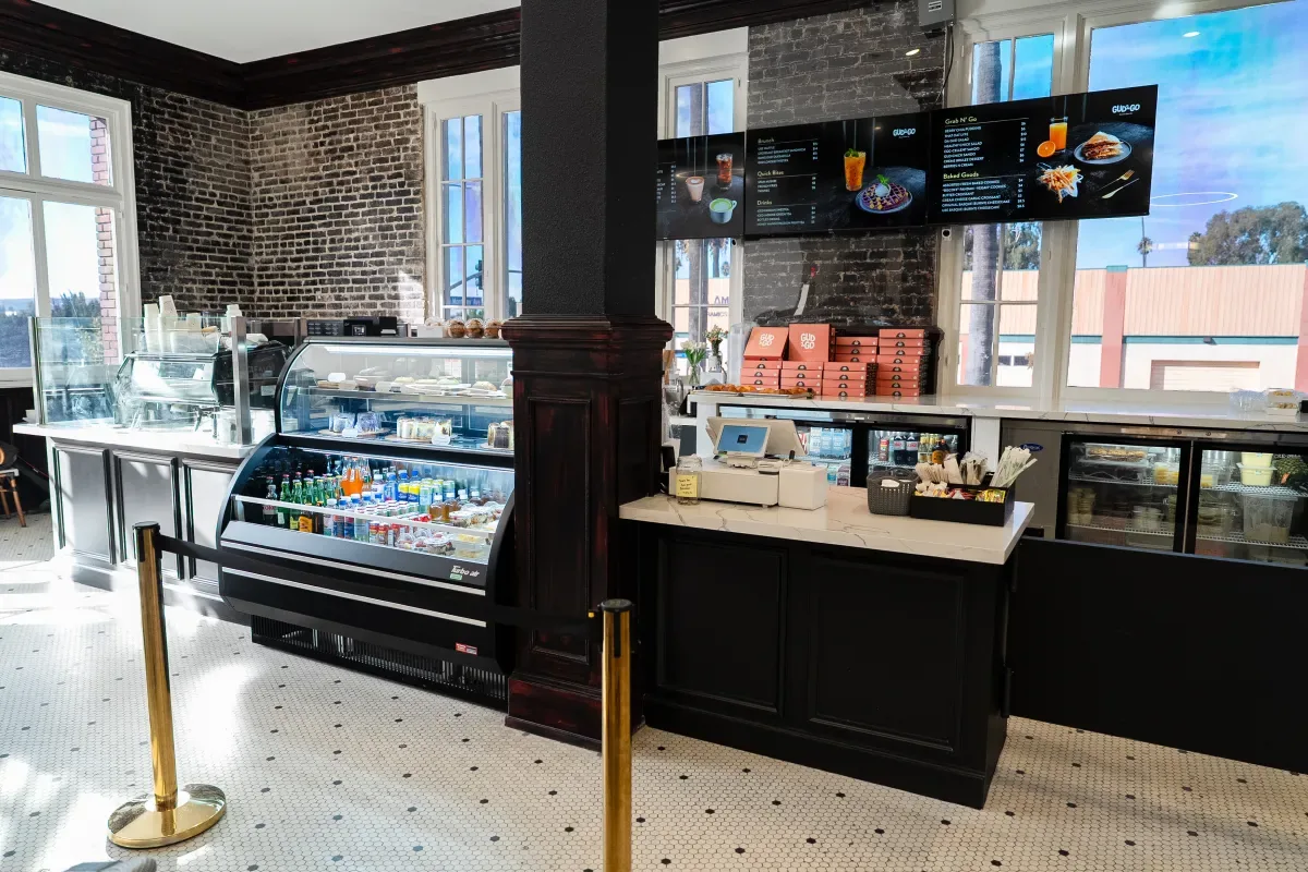 Order counter inside Take Ur Seat Cafe in Pomona — pastry case, digital menu boards, and exposed brick walls.